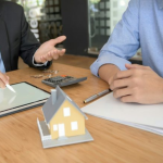 Two business people discuss plans with a small house model, tablet, and calculator on the table.