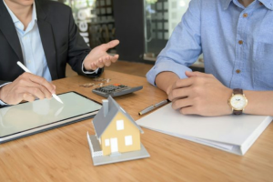 Two business people discuss plans with a small house model, tablet, and calculator on the table.